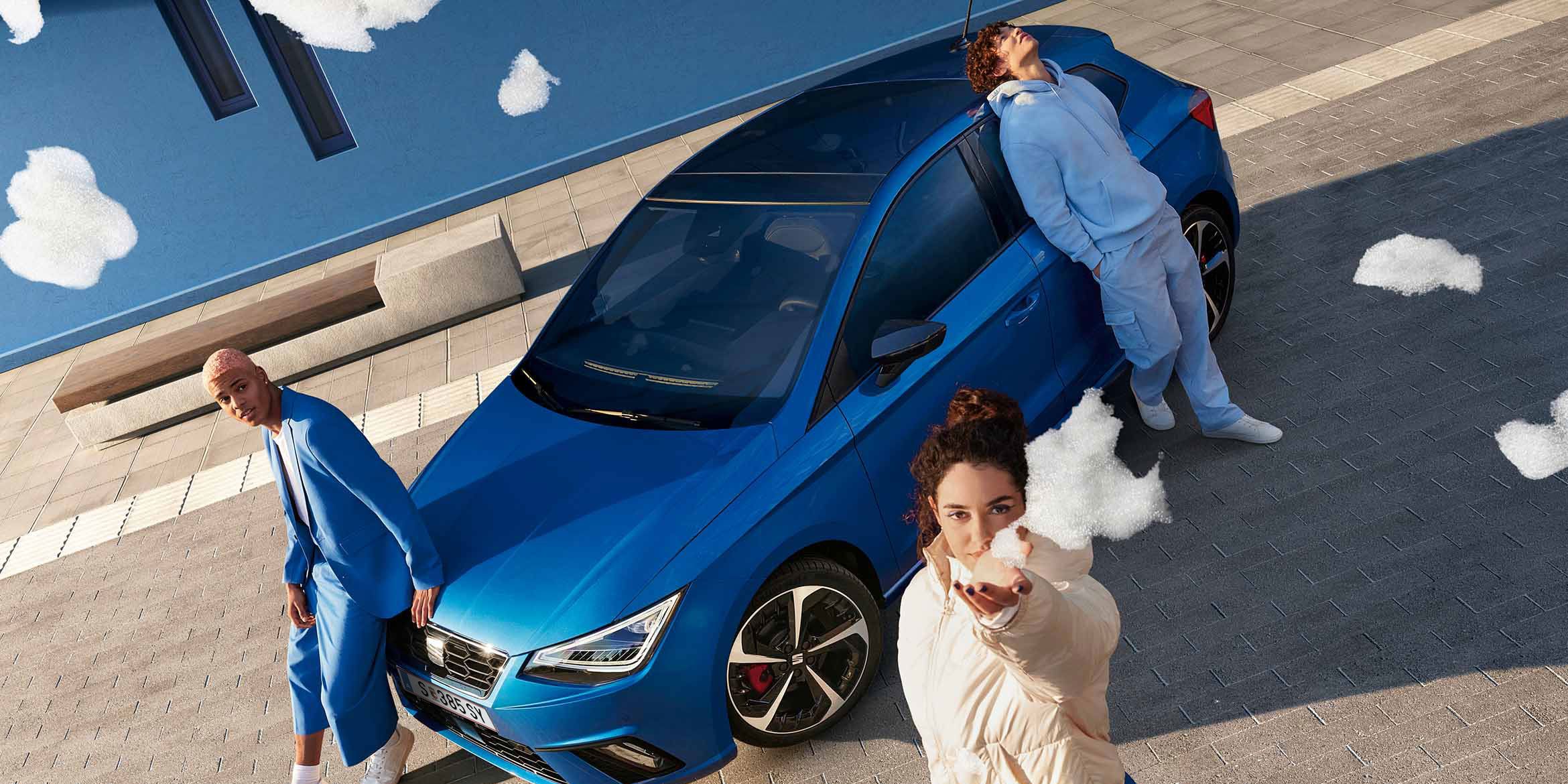 Ein blauer SEAT Ibiza aus der Vogelperspektive: Drei Personen, alle in blauer Kleidung, posieren am Auto – zwei lehnen an der Motorhaube, eine an der Seitentür. Eine der Personen hält Schaum in die Kamera
