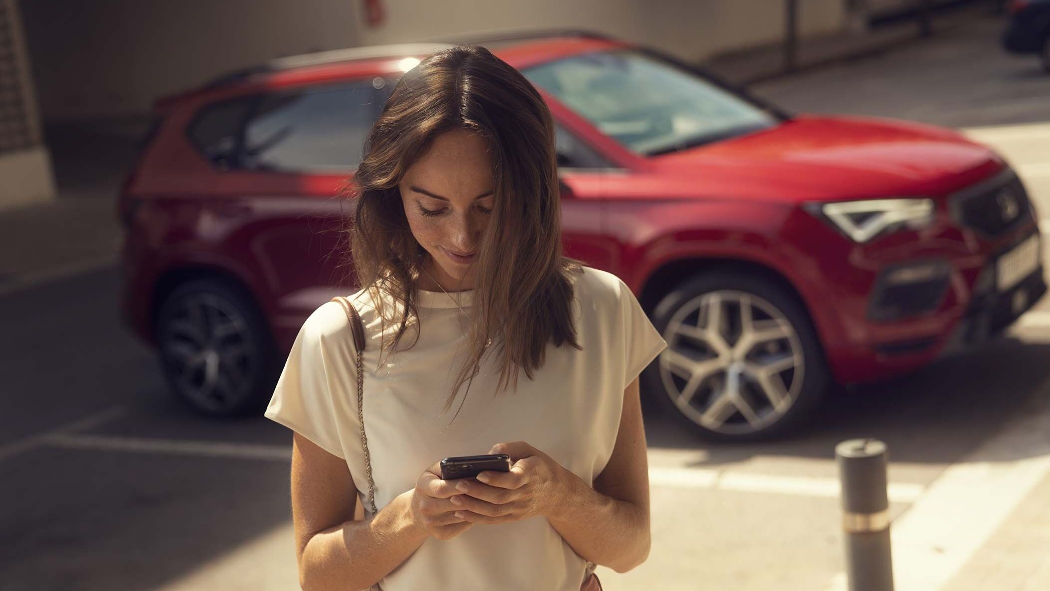 Eine Frau hält ein Smartphone in den Händen. Im Hintergrund steht ein roter SEAT.