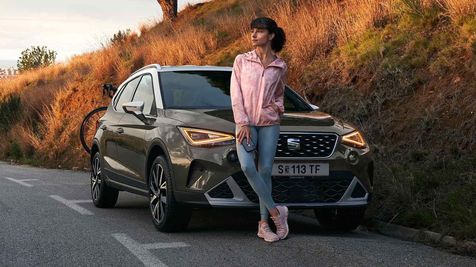 Eine Frau stützt sich auf die Motorhaube eines SEAT Arona in der Farbe Cliff Grey