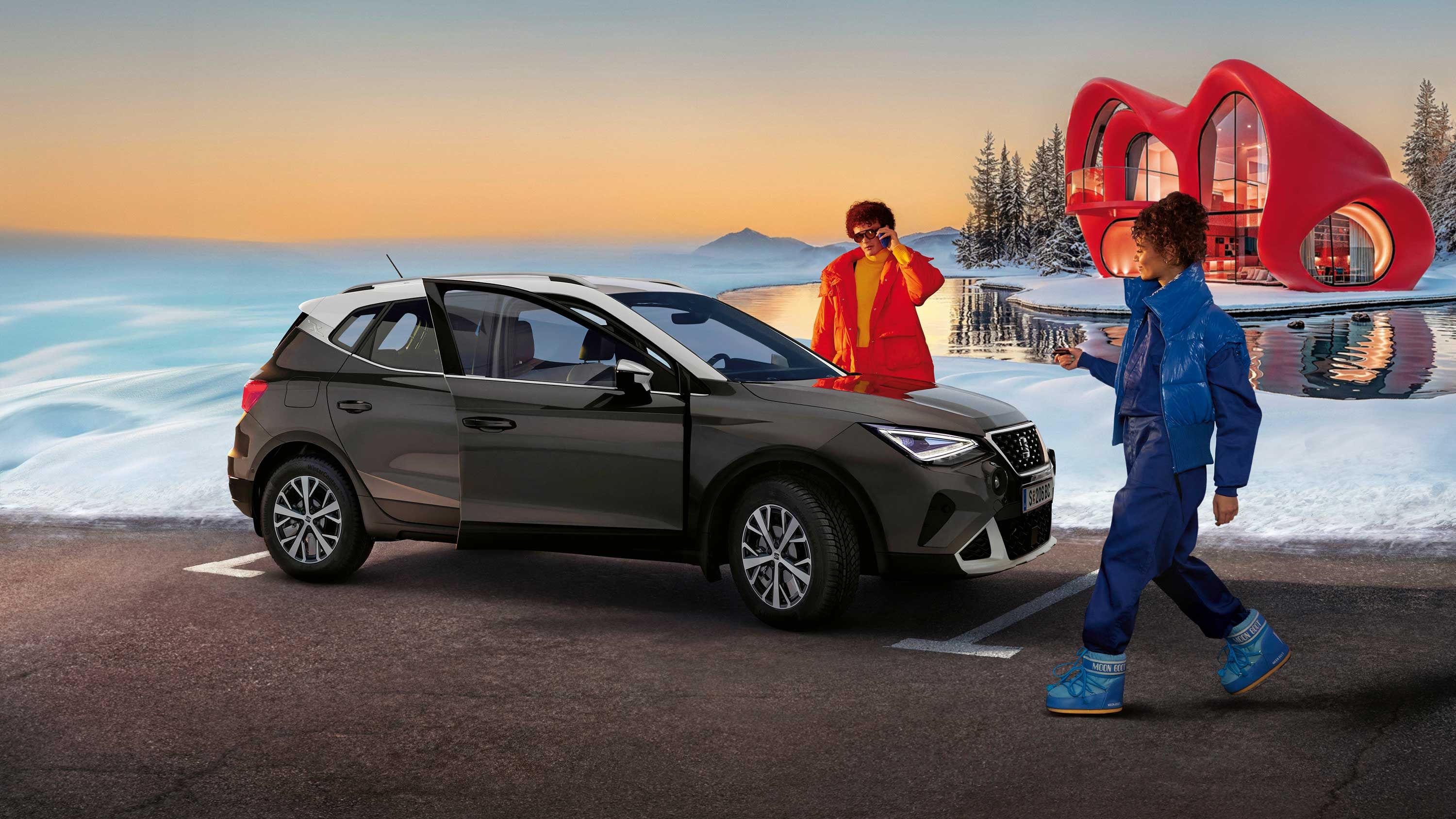 Eine Person steht neben einem SEAT Arona, eine weitere nähert sich dem Fahrzeug. Im Hintergrund sind Schnee, ein Haus und ein Sonnenunterganghimmel zu sehen