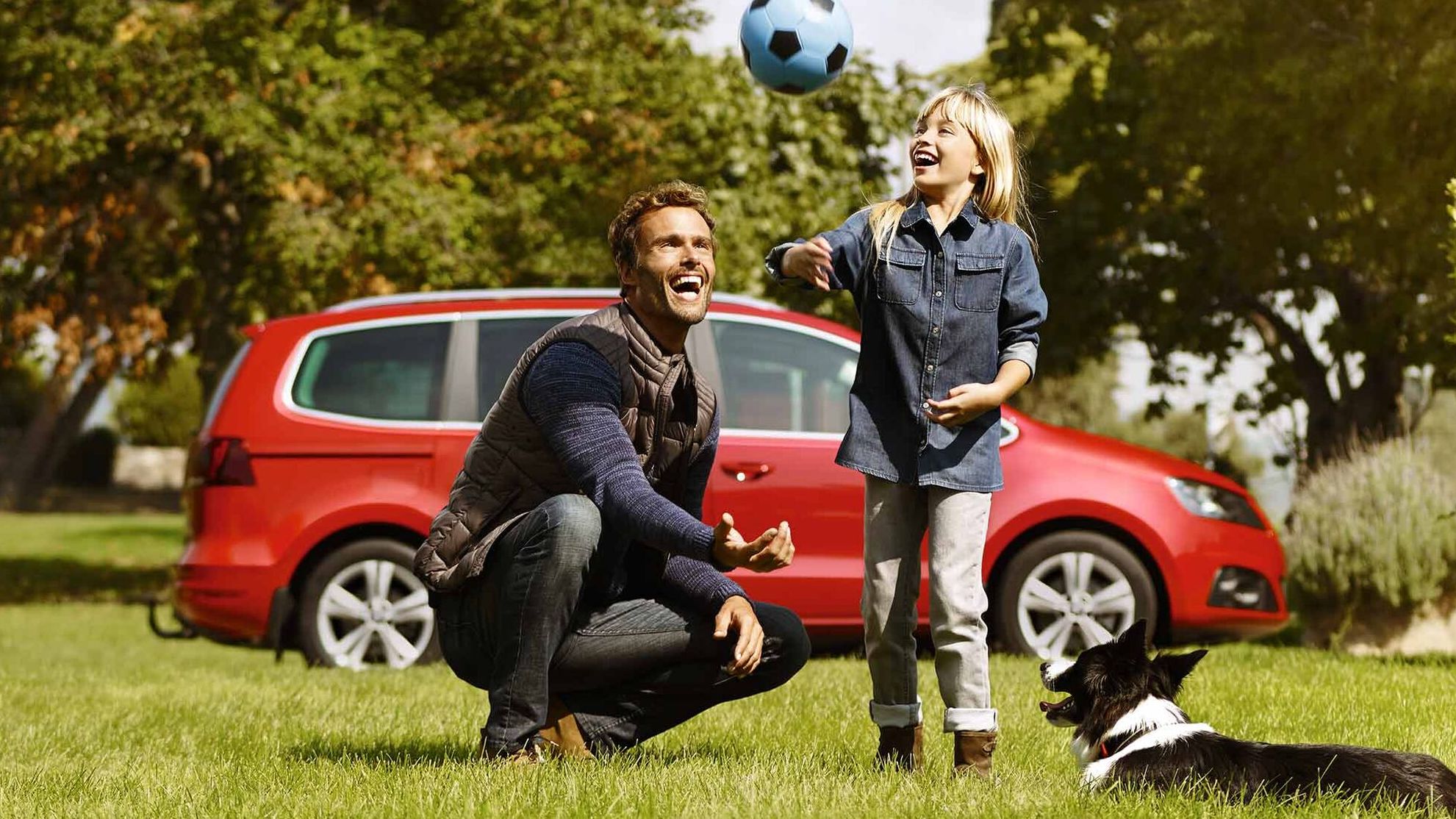 Im Hintergrund parkt ein roter SEAT Alhambra, während ein Vater gemeinsam mit seiner Tochter und ihrem Hund auf einer Wiese Ball spielt