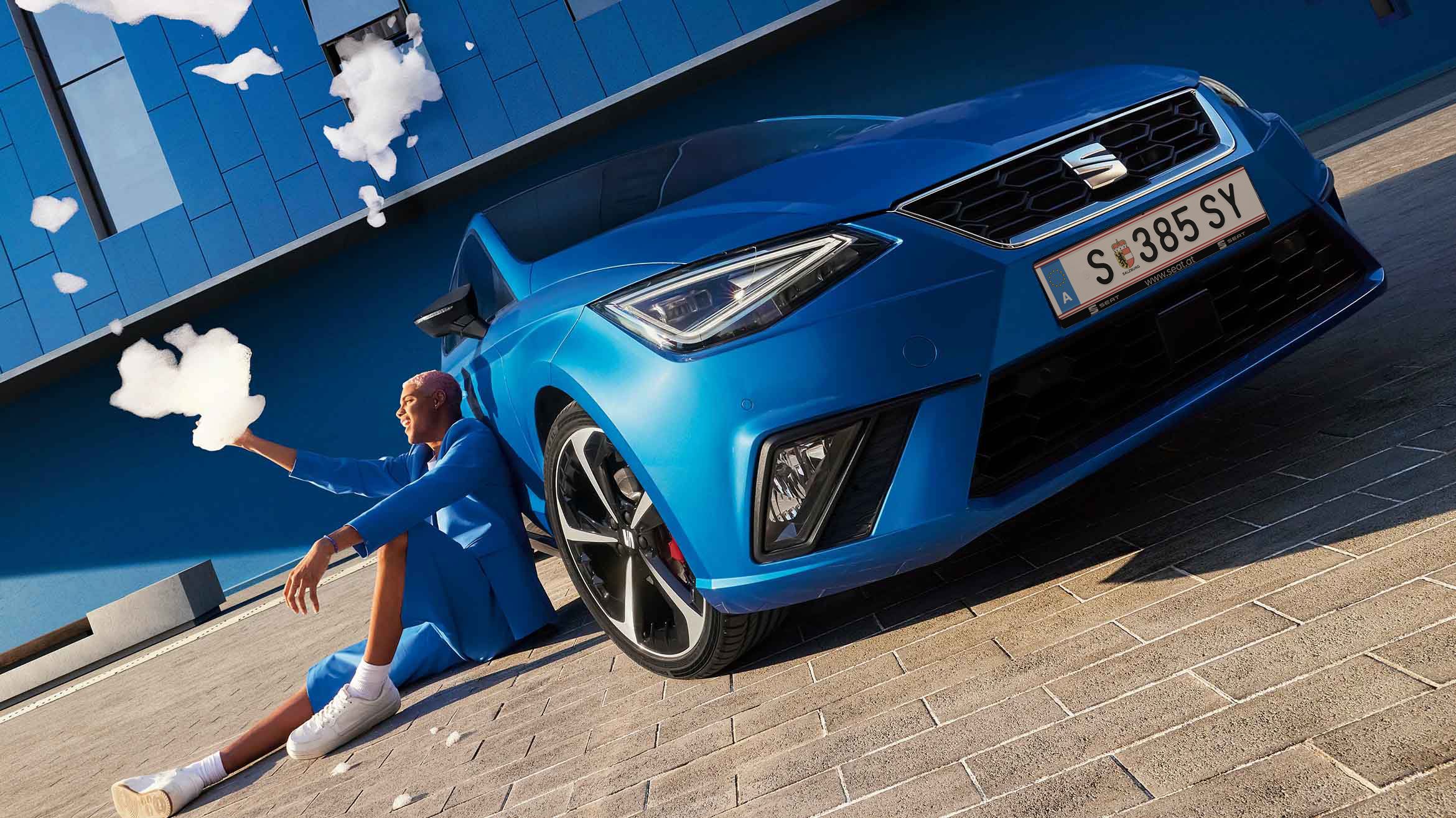 Ein blauer SEAT Ibiza steht vor einem ebenfalls blauen Gebäude. Davor sitzt ein Mann in einem blauen Anzug mit dem Rücken zu seinem Auto und wirft Seifenschaum in die Luft
