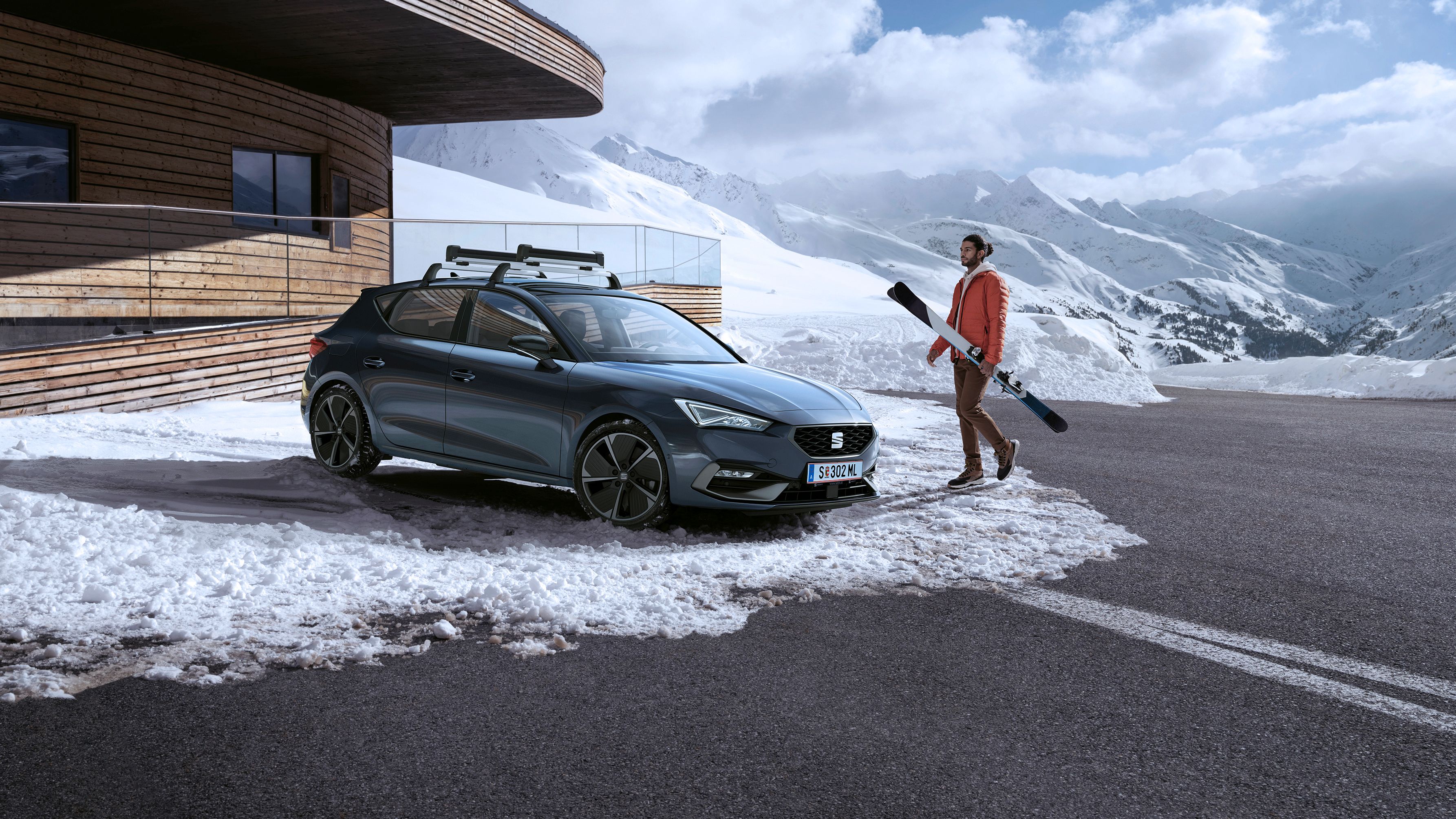 Ein Mann trägt Ski zu einem grauen SEAT Leon, der vor einem Haus in den verschneiten Bergen geparkt ist.