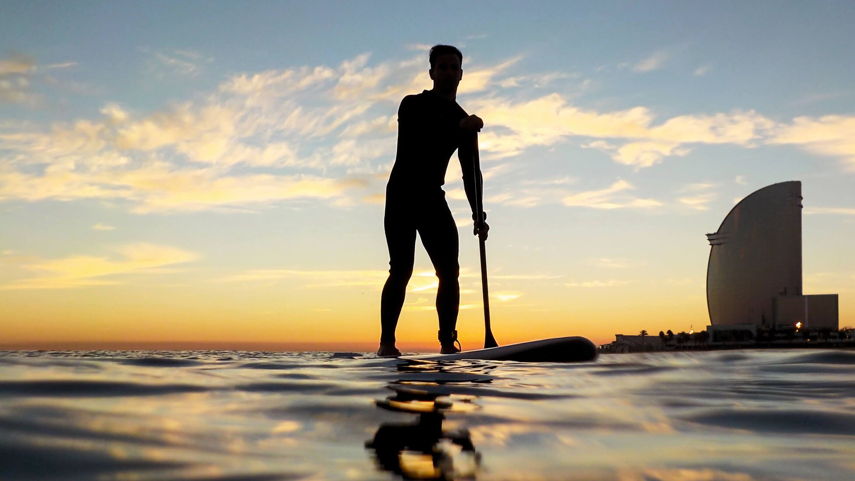 Silhouette eines Mannes beim Paddle-Surfen am Strand von Barceloneta, mit dem markanten W Hotel im Hintergrund