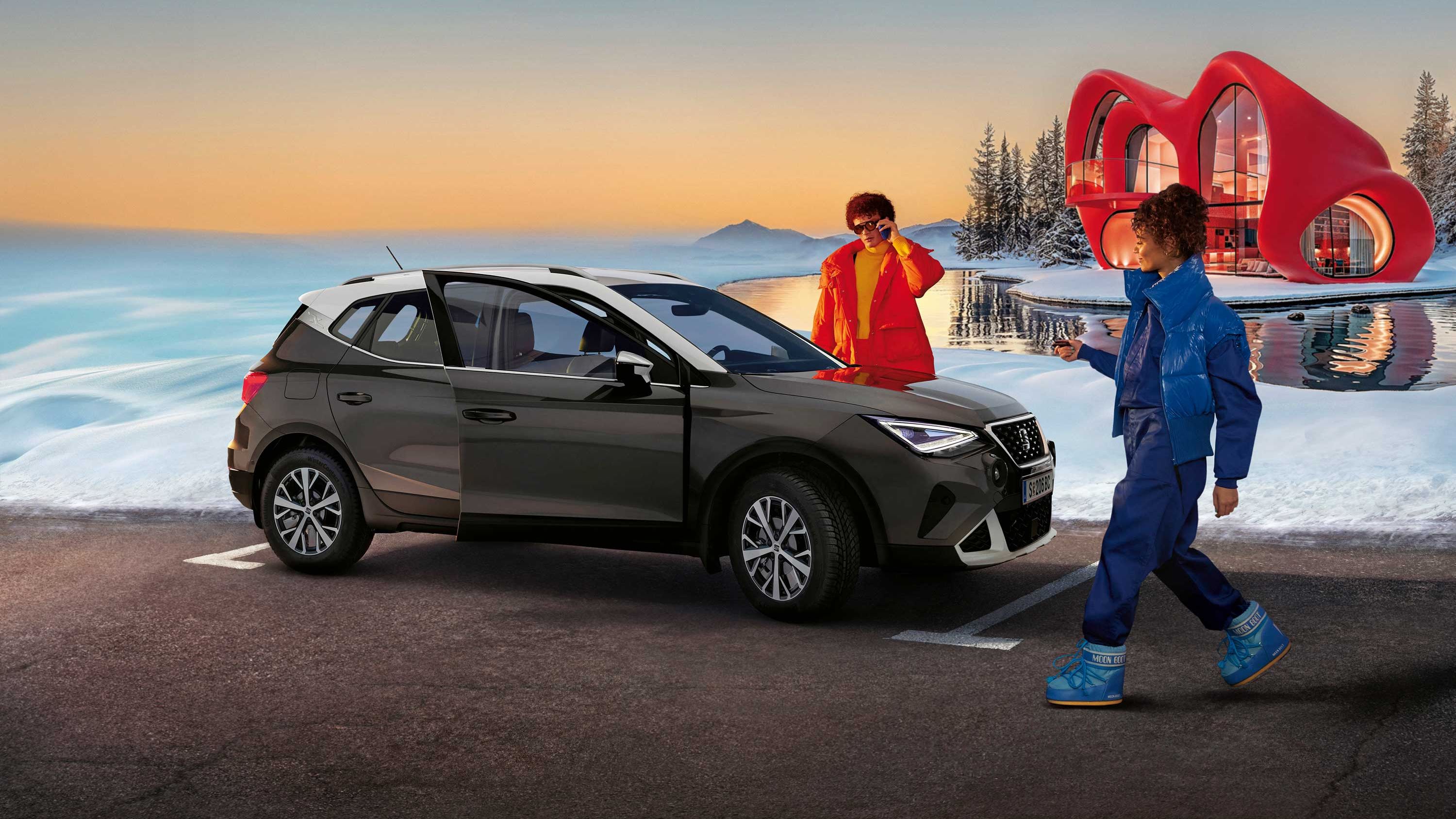 Eine Person steht neben einem SEAT Arona, eine weitere nähert sich dem Fahrzeug. Im Hintergrund sind Schnee, ein Haus und ein Sonnenunterganghimmel zu sehen