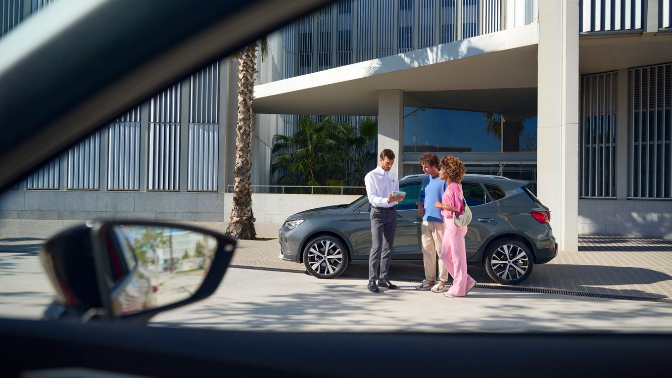 Drei Personen stehen vor einem grauen SEAT Arona im Gespräch: Ein Paar erkundigt sich interessiert, während ein Mann ihnen etwas zeigt