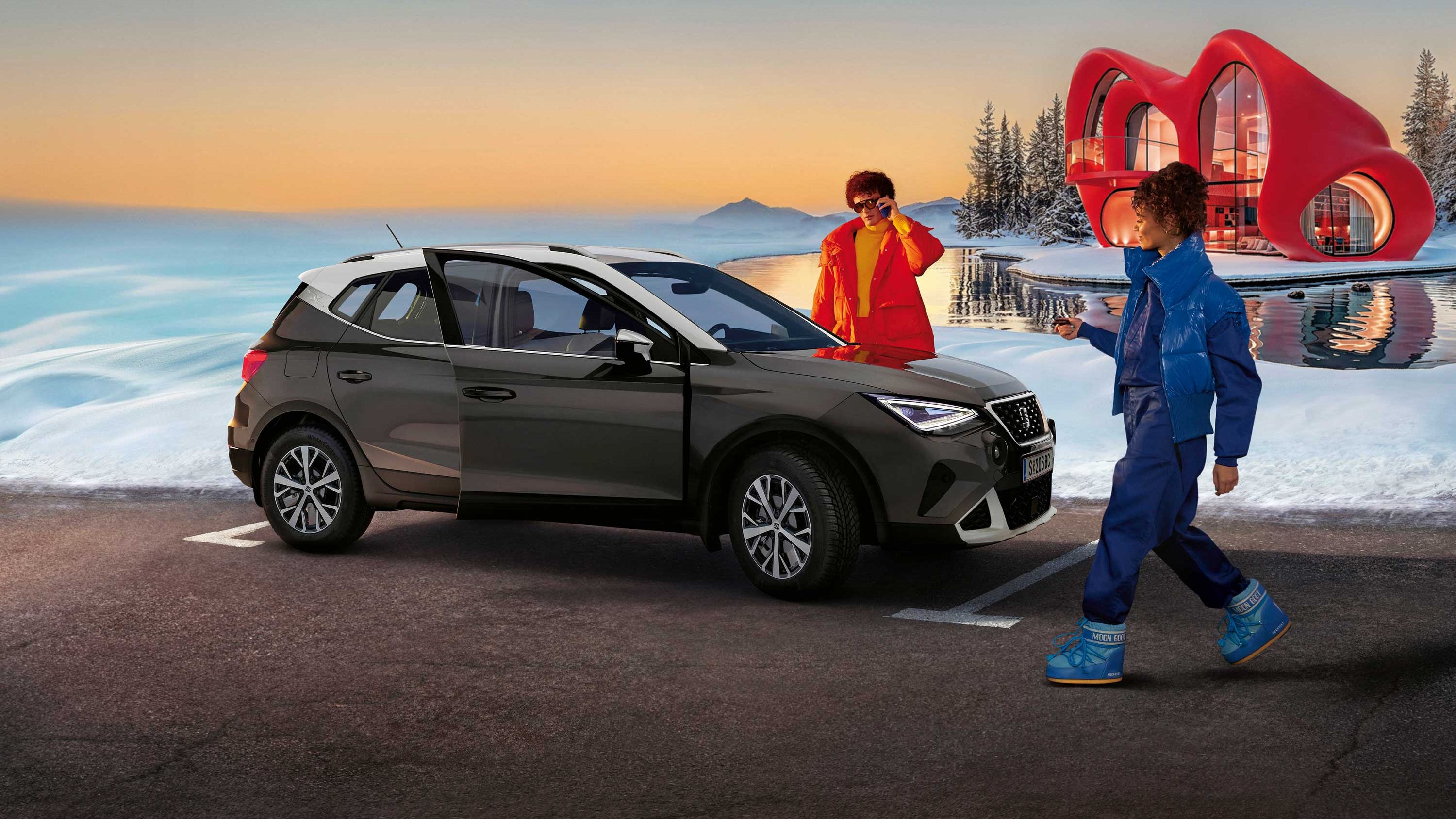 Eine Person steht neben einem SEAT Arona, eine weitere nähert sich dem Fahrzeug. Im Hintergrund sind Schnee, ein Haus und ein Sonnenunterganghimmel zu sehen
