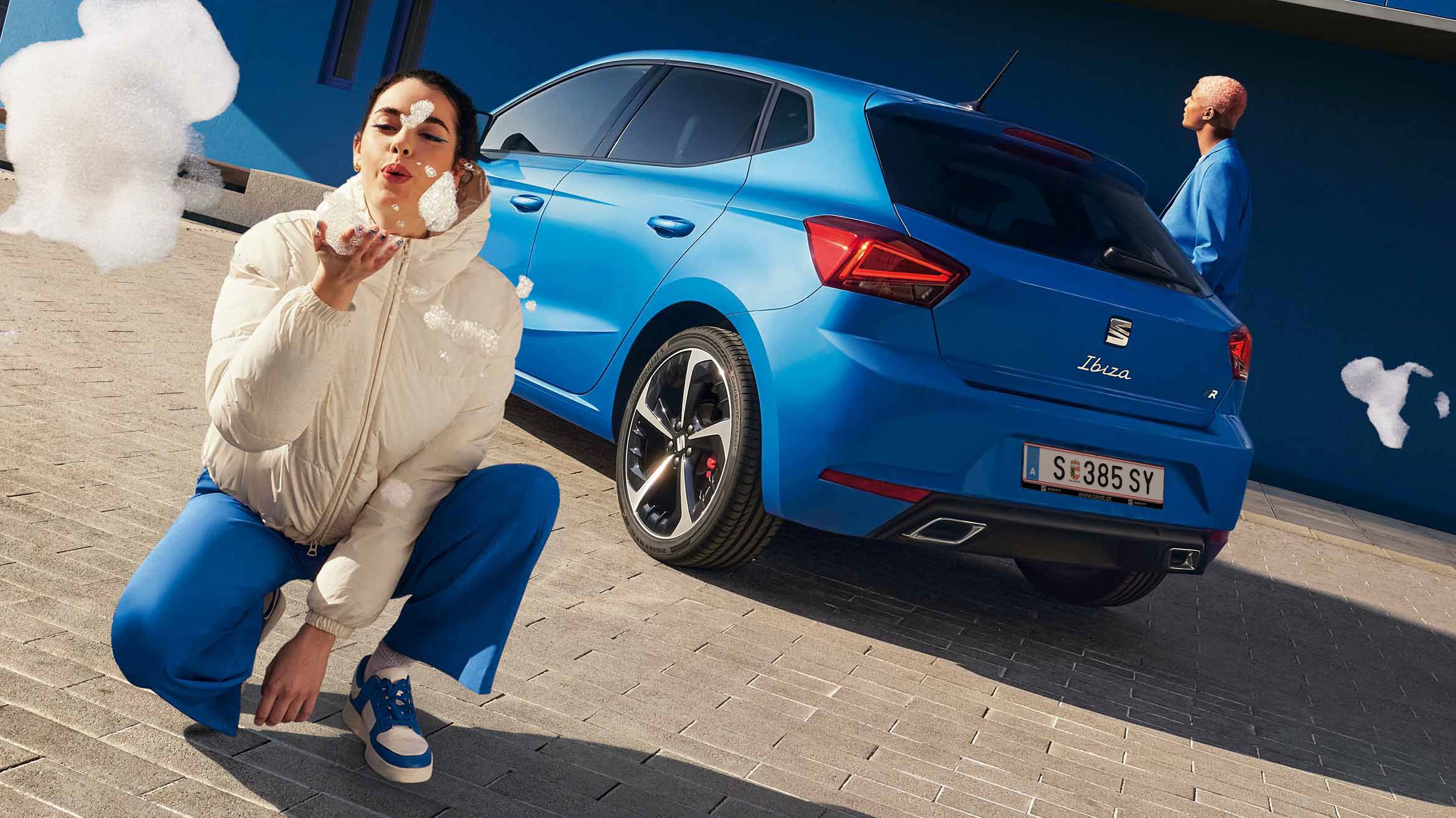 Ein blauer SEAT Ibiza steht mit dem Heck zu einem blauen Gebäude. Ein Mann im blauen Anzug geht daran vorbei, während eine Frau direkt vor der Kamera kniet und Seifenblasen in Richtung Linse pustet
