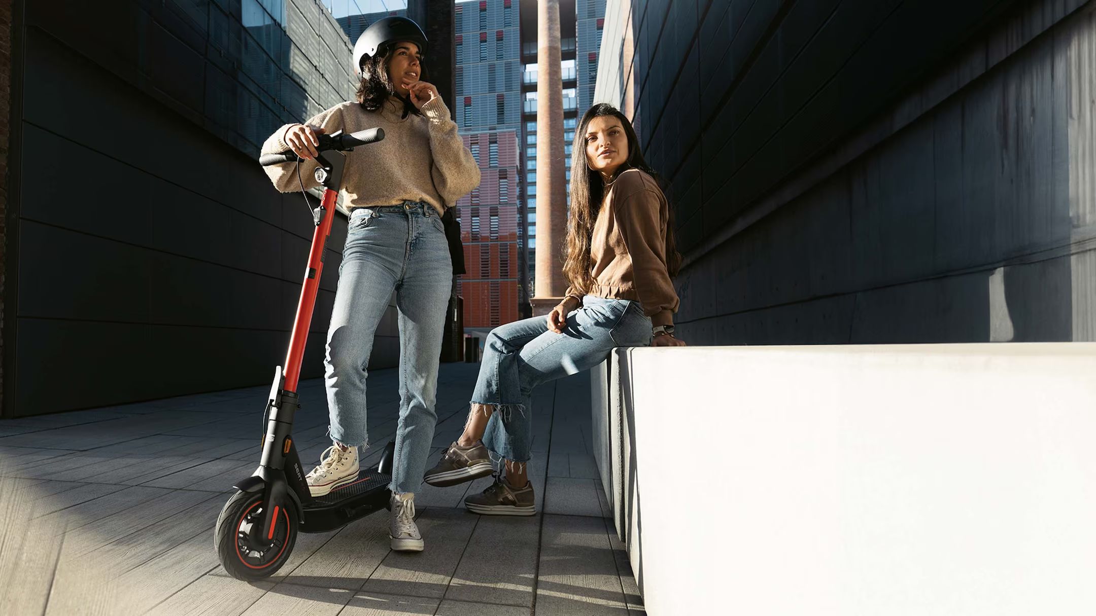 Eine Frau steht mit einem Bein auf ihren SEAT MO 65 Kickscooter, während eine weitere Frau entspannt auf einer Hausmauer sitzt
