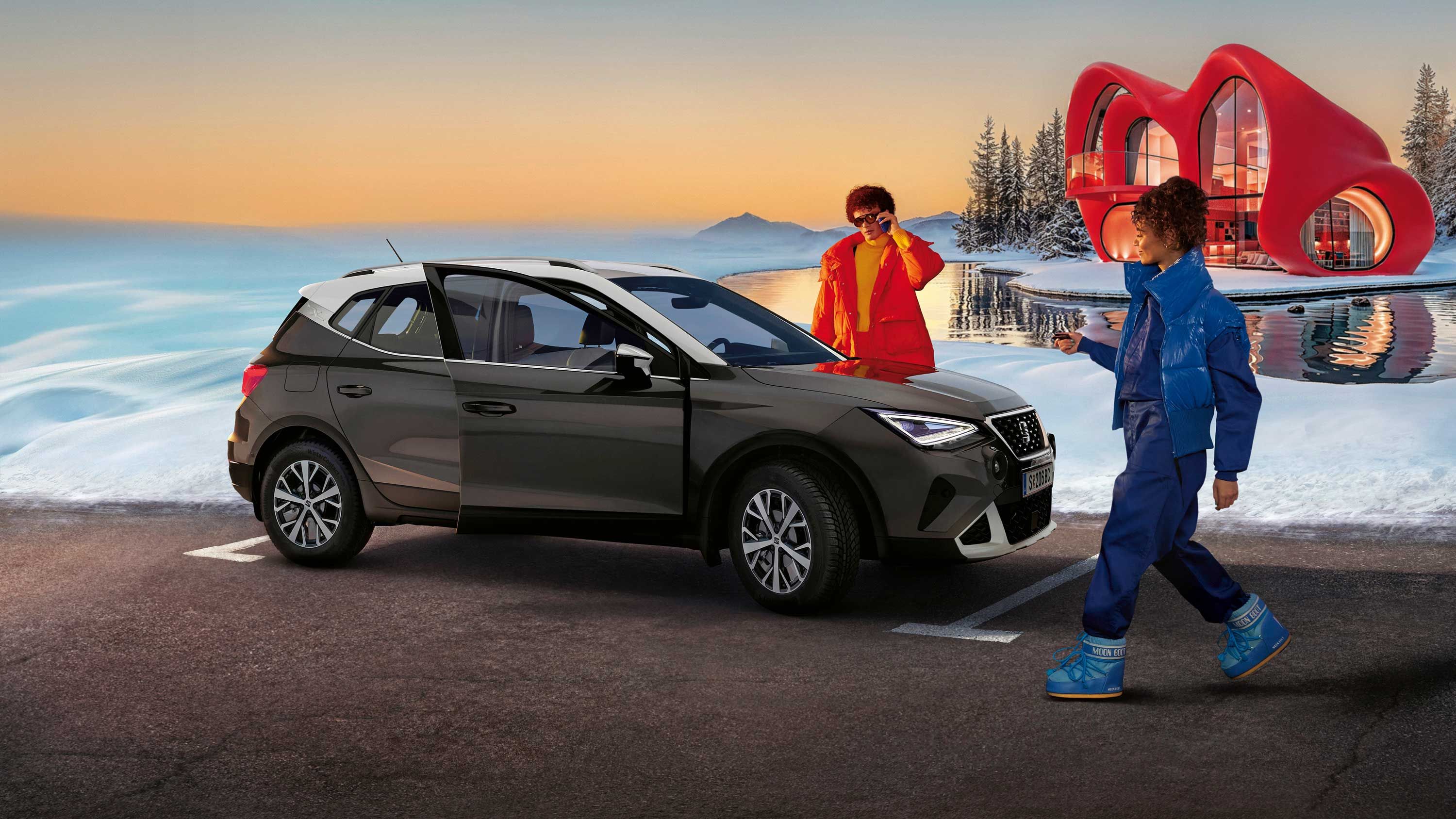 Eine Person steht neben einem SEAT Arona, eine weitere nähert sich dem Fahrzeug. Im Hintergrund sind Schnee, ein Haus und ein Sonnenunterganghimmel zu sehen