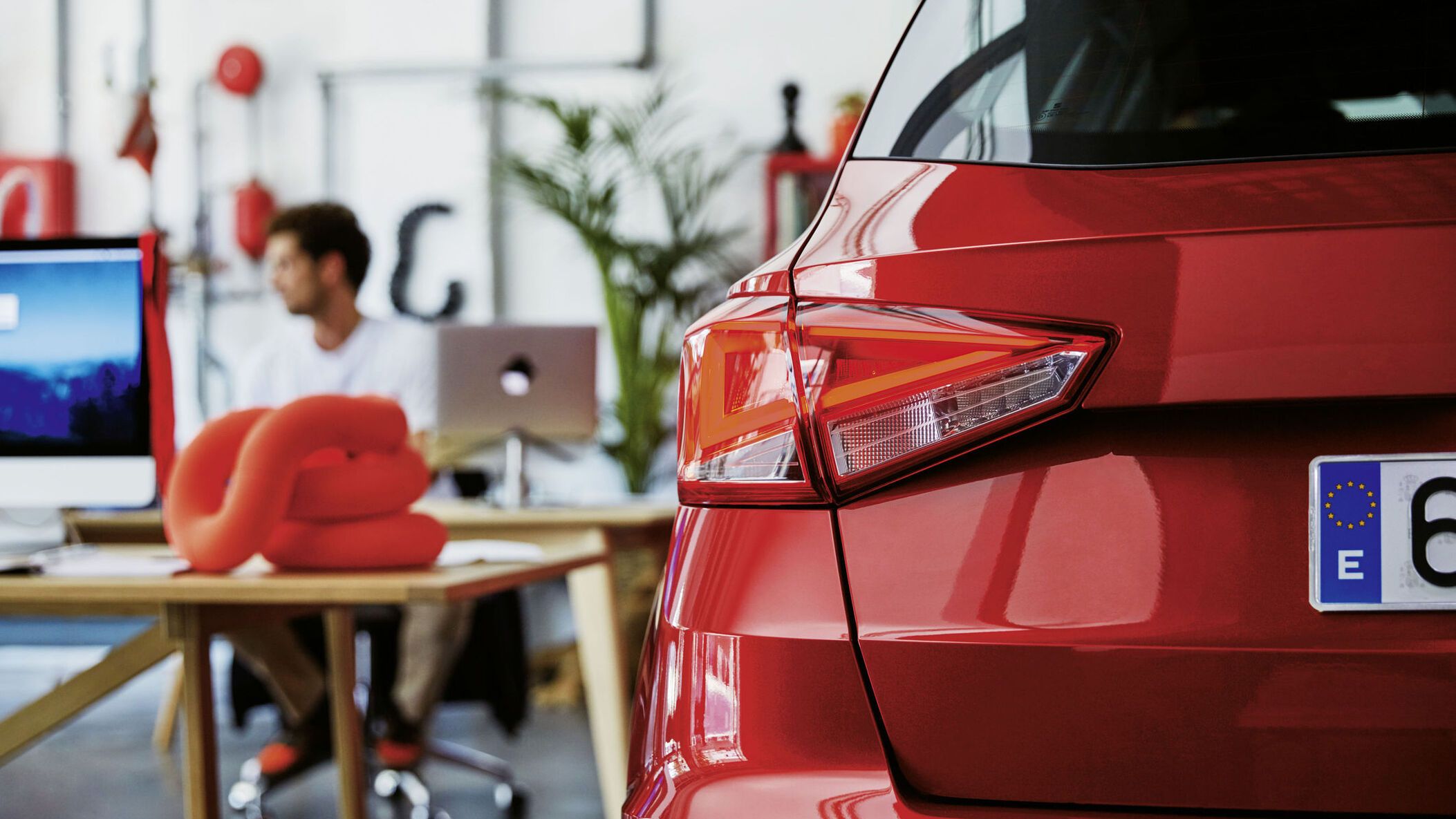 Pohľad na časť zadných partií  modelu SEAT Arona, ktorý je zaparkovaný v showroome, pozadí vidieť stoly za jedným sedí predajca a pracuje.