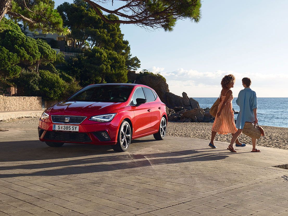 Ein roter SEAT Ibiza steht neben einem Strand und zwei Freundinnen gehen gerade Richtung Meer 