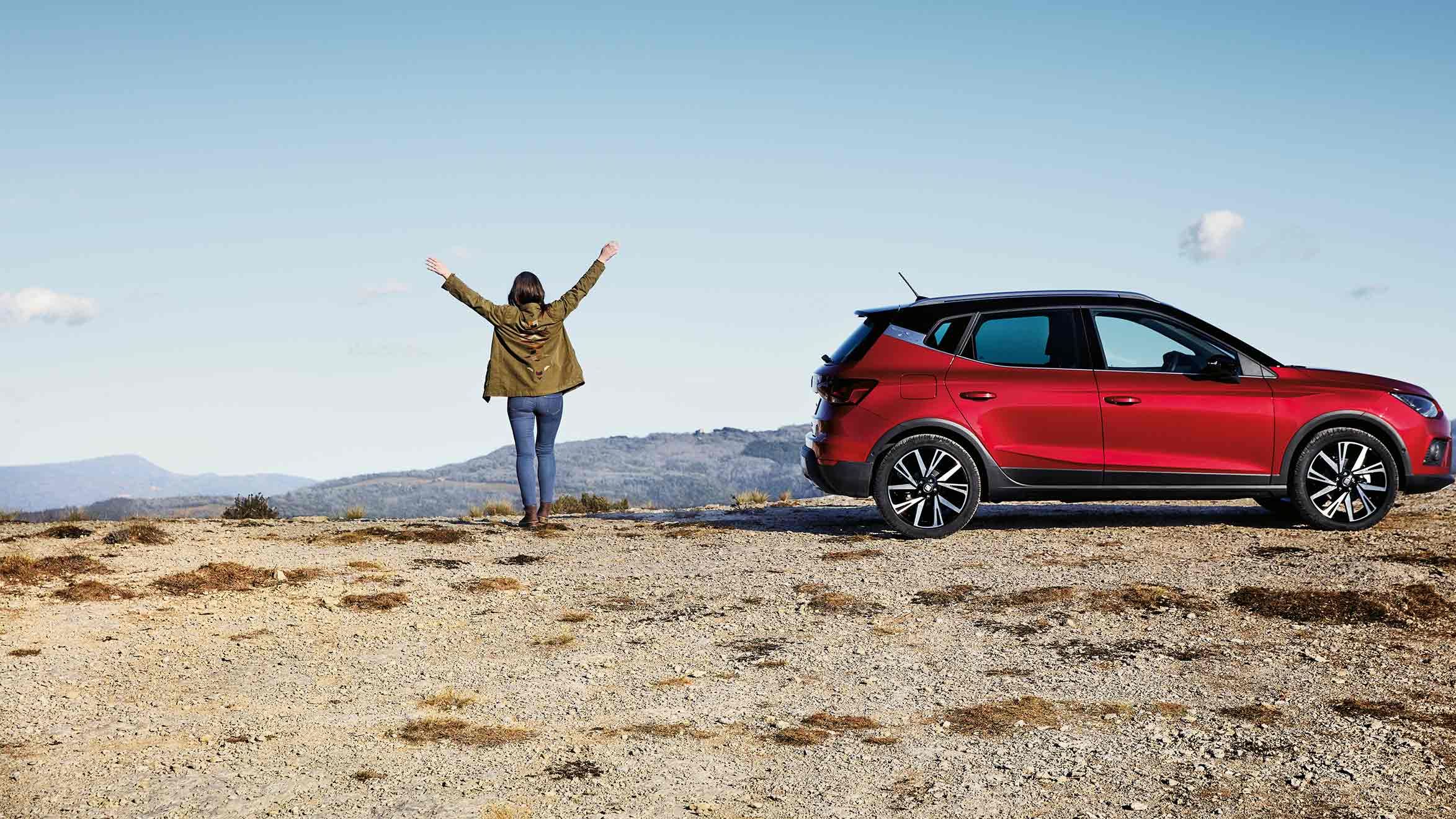 Eine Frau steht neben einem roten SEAT Arona auf einer Bergklippe und streckt die Arme triumphierend in die Höhe, während sie die Aussicht genießt