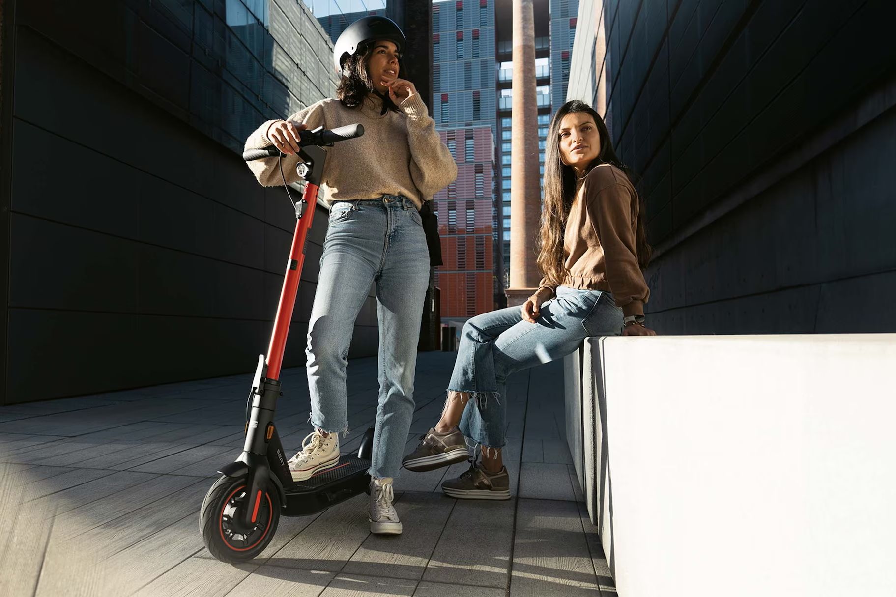 Eine Frau steht mit einem Bein auf ihren SEAT MO 65 Kickscooter, während eine weitere Frau entspannt auf einer Hausmauer sitzt