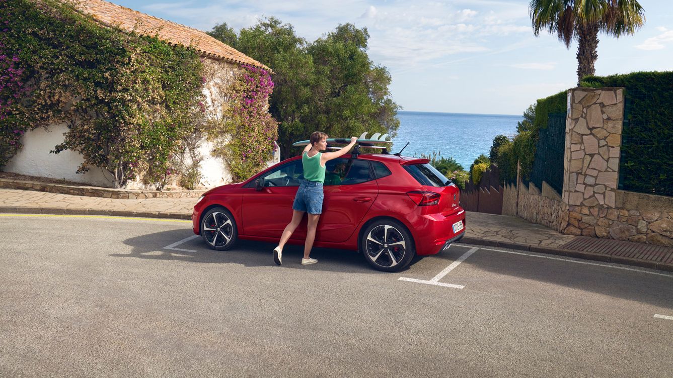 Ein roter SEAT Ibiza steht am Straßenrand, während eine Frau das Surfbrett vom Dach abnimmt