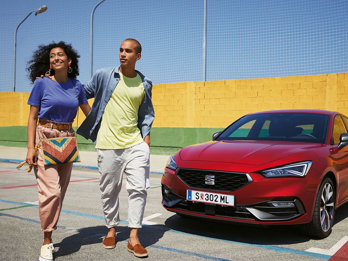 Eine Frau und ein Mann gehen neben einem geparkten SEAT Leon FR in Desire Red
