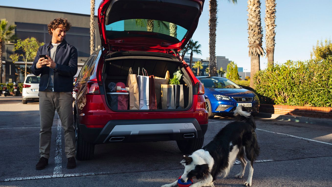 Ein Mann hat seine Einkäufe im SEAT Arona verstaut, während ein Hund vor dem Auto spielt