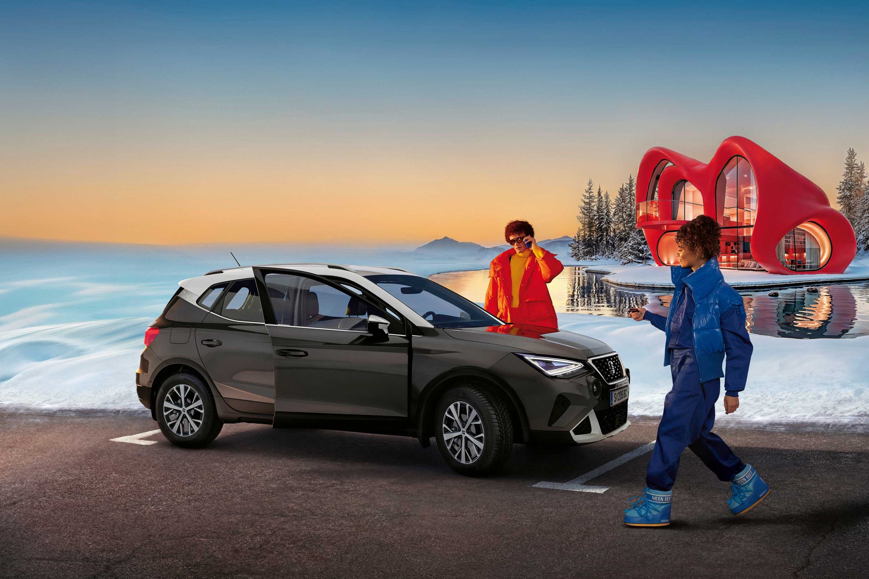 Eine Person steht neben einem SEAT Arona, eine weitere nähert sich dem Fahrzeug. Im Hintergrund sind Schnee, ein Haus und ein Sonnenunterganghimmel zu sehen