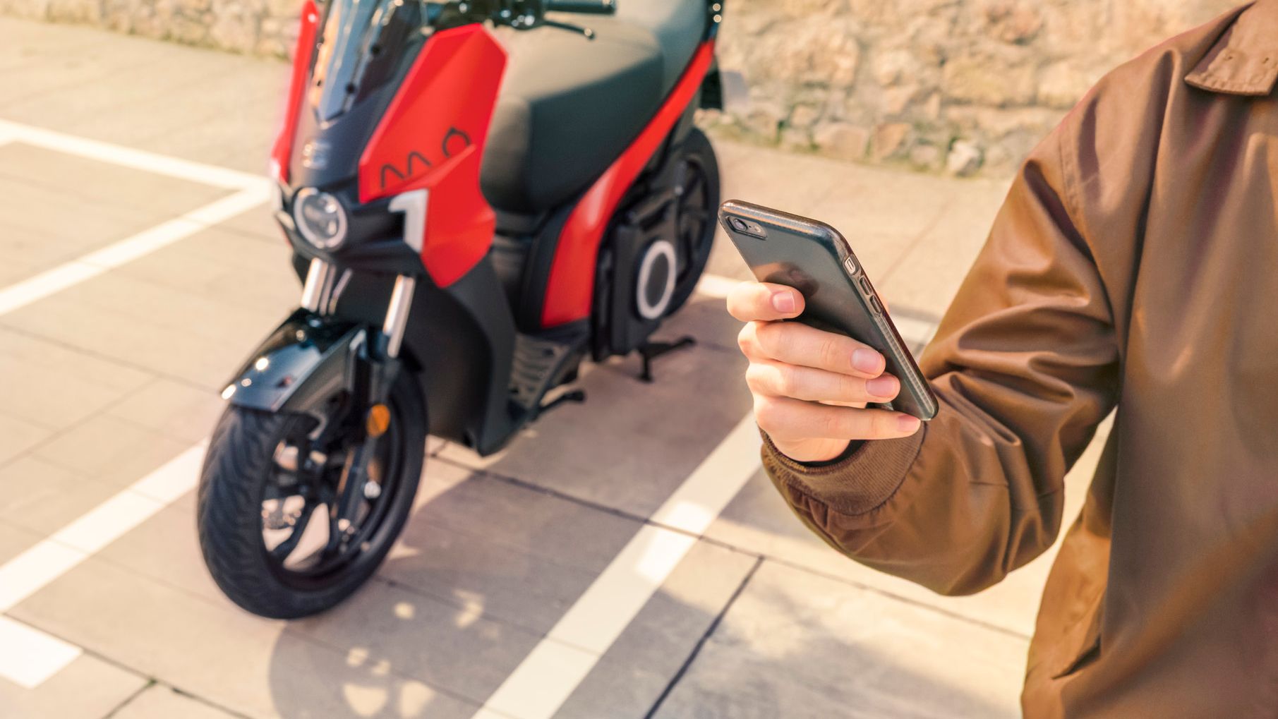 Eine Person hält ein Smartphone in der Hand, im Hintergrund steht ein SEAT MO 125.