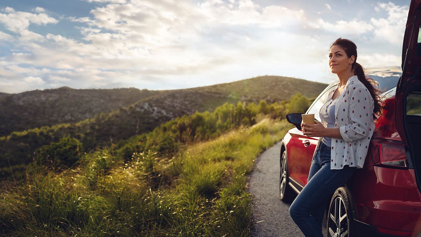 Eine Frau lehnt an einem roten SEAT Ibiza, hält einen Becher in der Hand und blickt auf die umliegende Berglandschaft entlang einer Landstraße