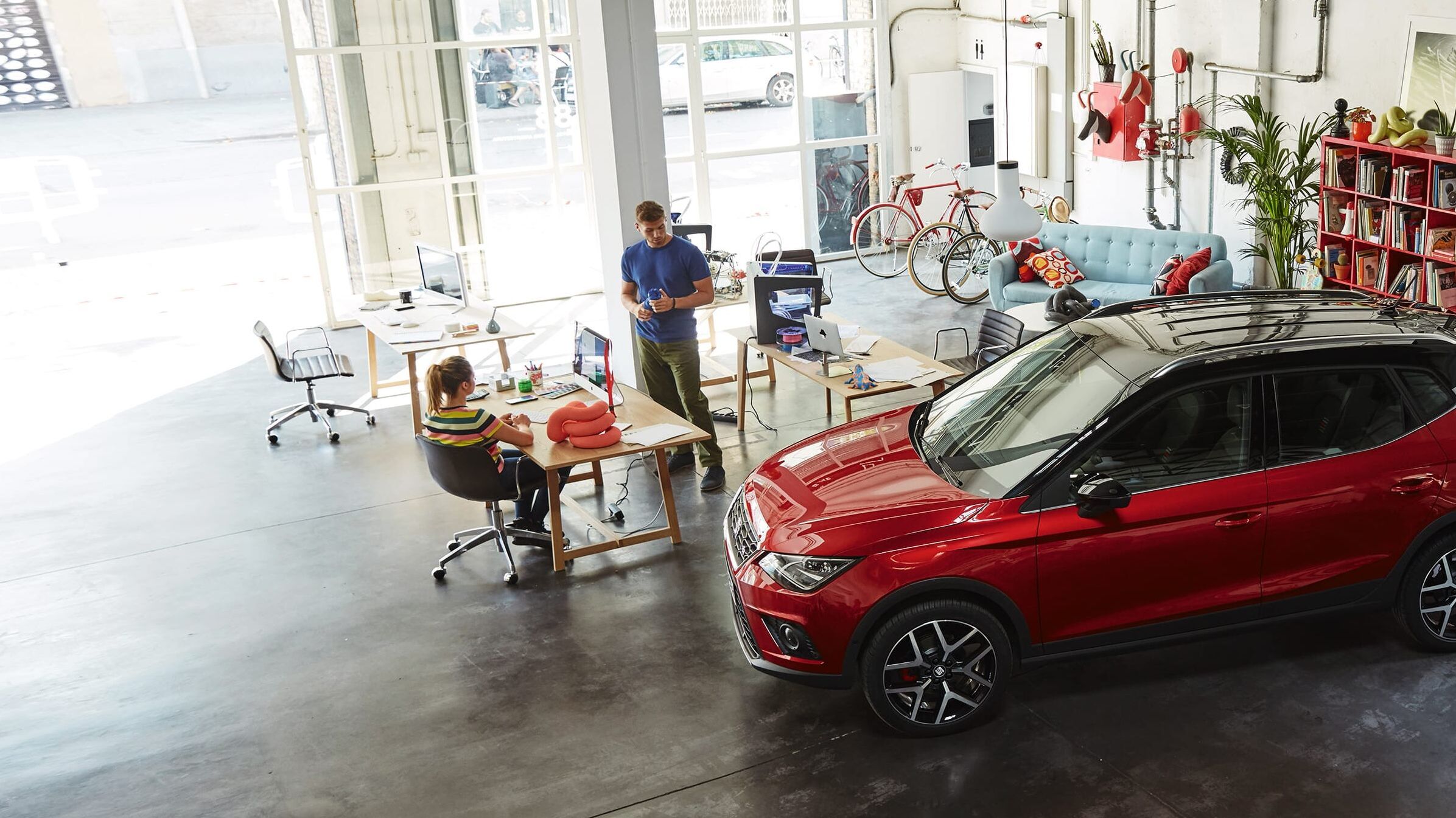 Ein roter SEAT Arona steht in einem modernen Büroraum, während sich zwei Mitarbeitende im Hintergrund unterhalten