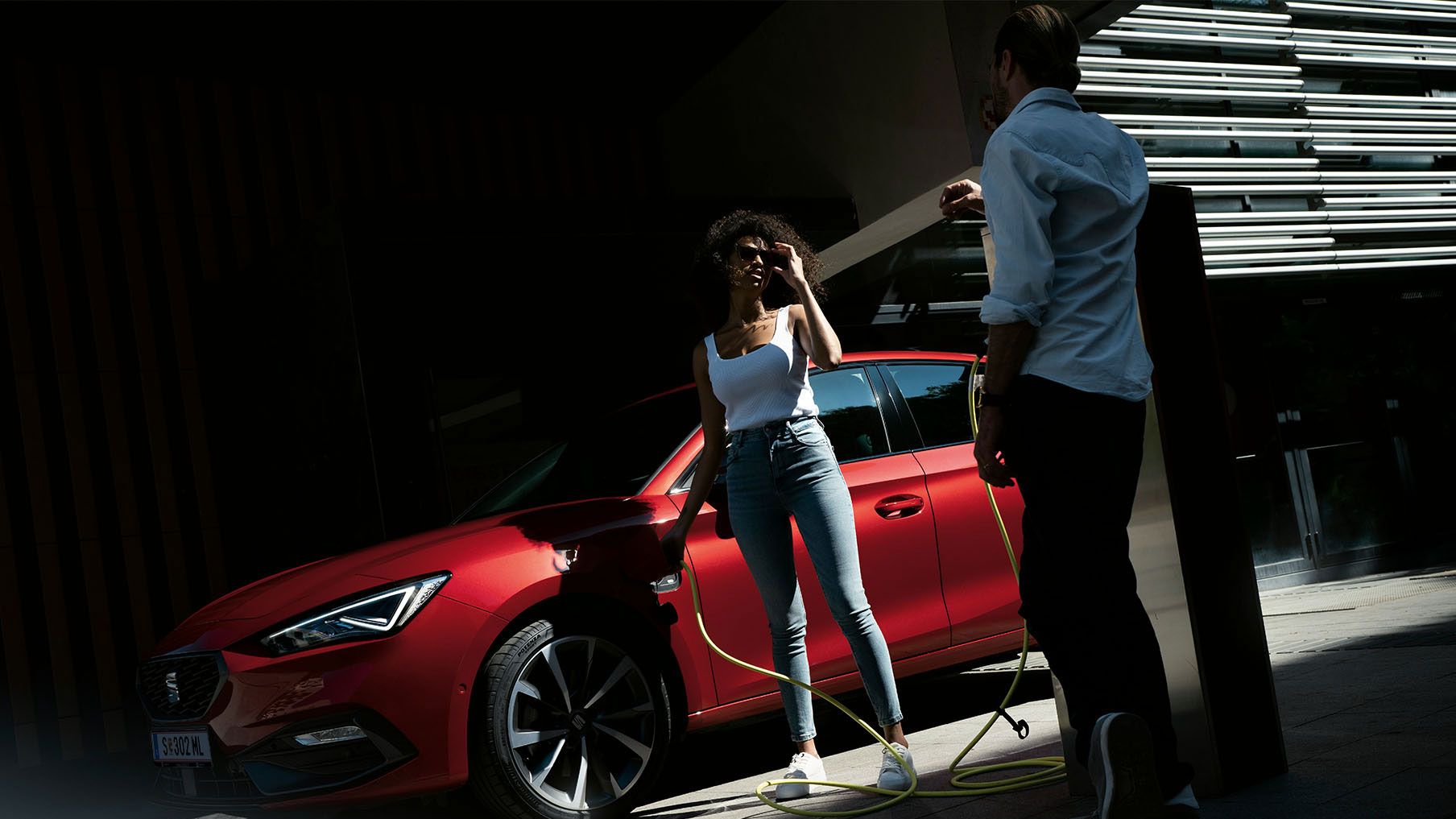 Eine Frau lädt gerade ihrem roten SEAT Leon an der Ladesäule und redet nebenbei mit einem Mann der bei der Ladesäule lehnt 