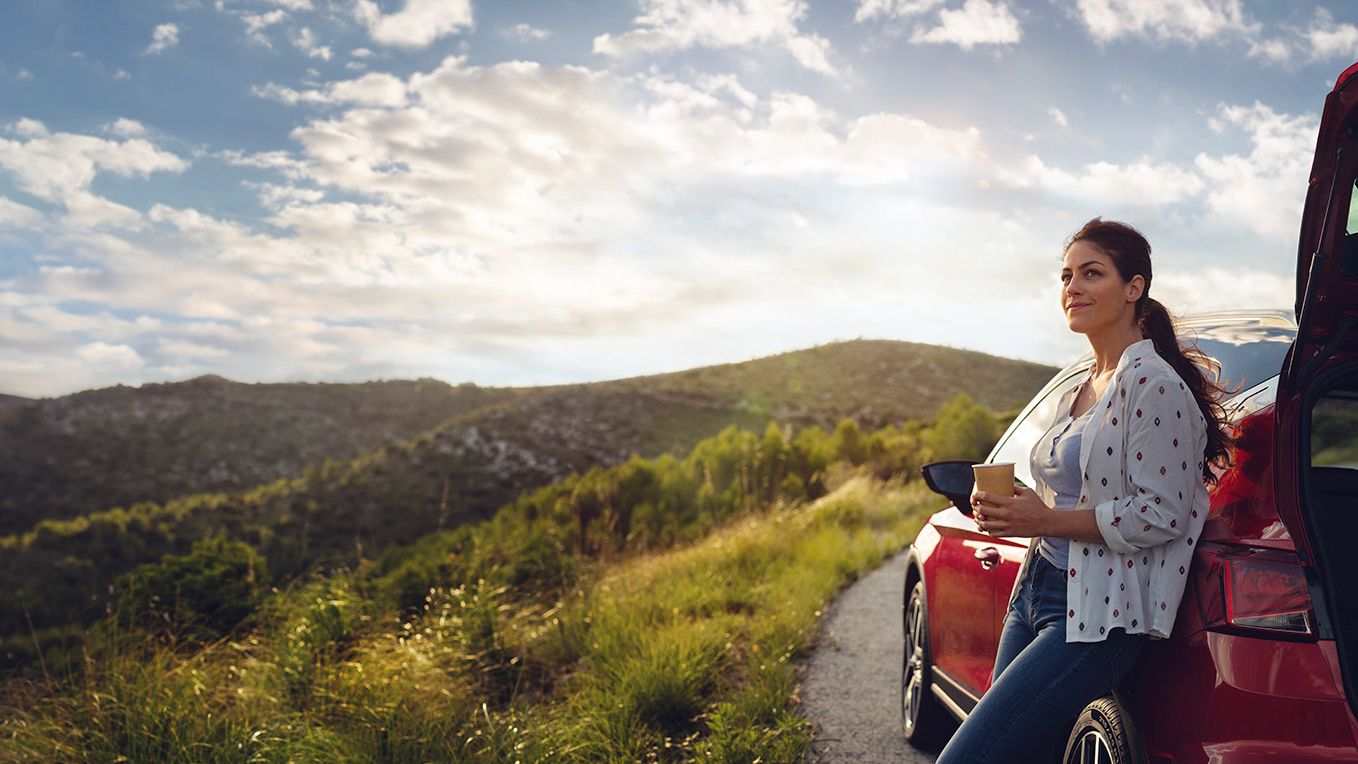 Eine Frau lehnt an einem roten SEAT Ibiza, hält einen Becher in der Hand und blickt auf die umliegende Berglandschaft entlang einer Landstraße.