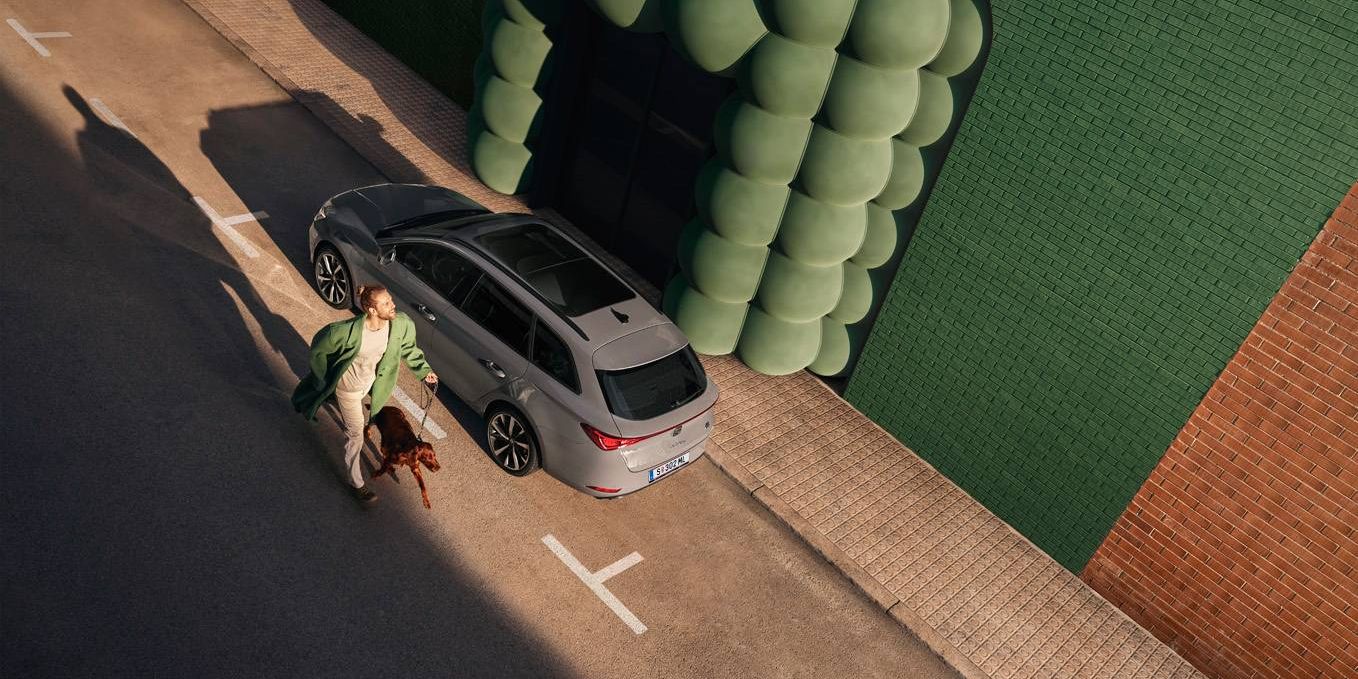 Blick von oben auf einen grauen SEAT Leon SP Kombi an dem ein Mann mit Hund vorbei marschiert.