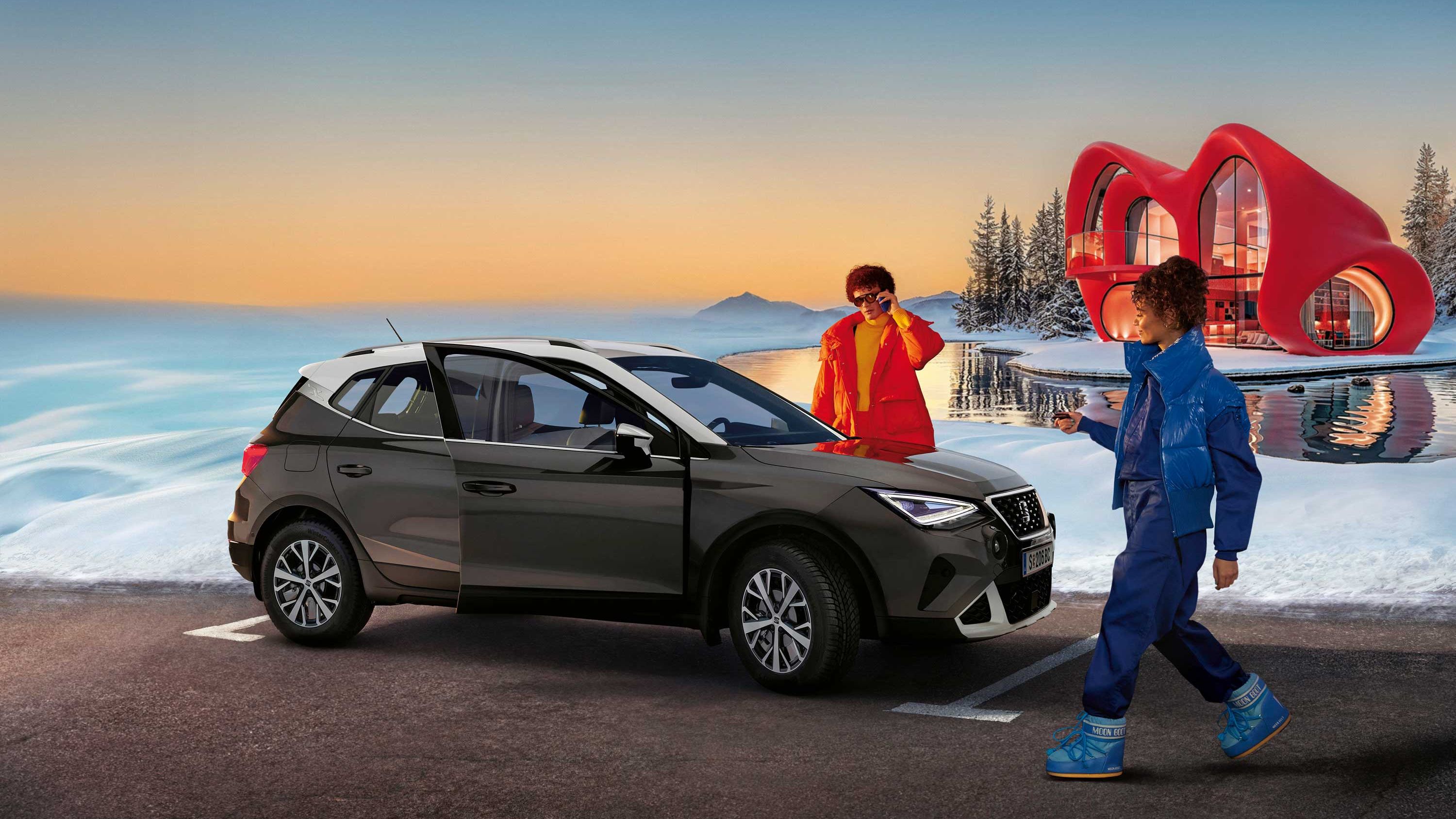 Eine Person steht neben einem SEAT Arona, eine weitere nähert sich dem Fahrzeug. Im Hintergrund sind Schnee, ein Haus und ein Sonnenunterganghimmel zu sehen