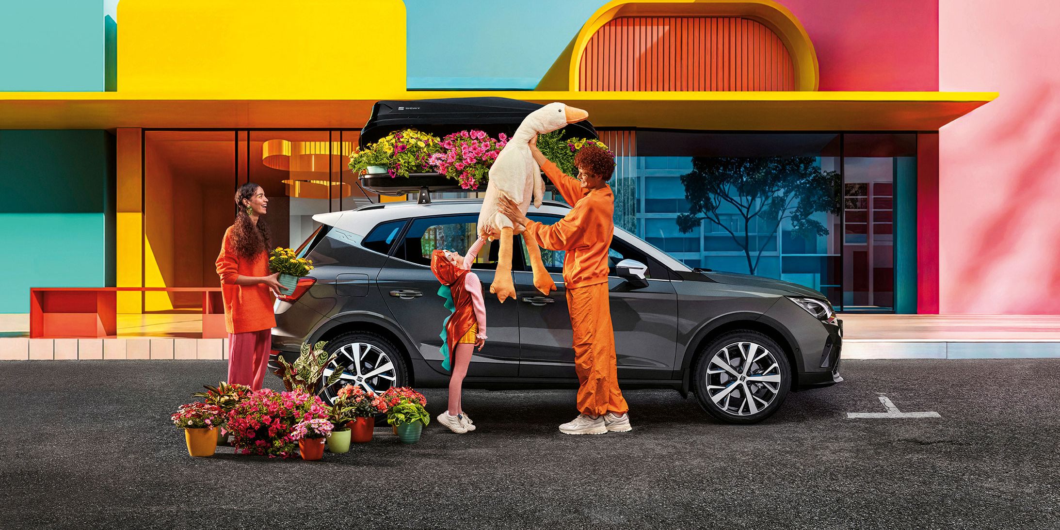 Eine Familie mit Blumen und einem Stofftier befindet sich vor einem SEAT Arona mit Dachbox.