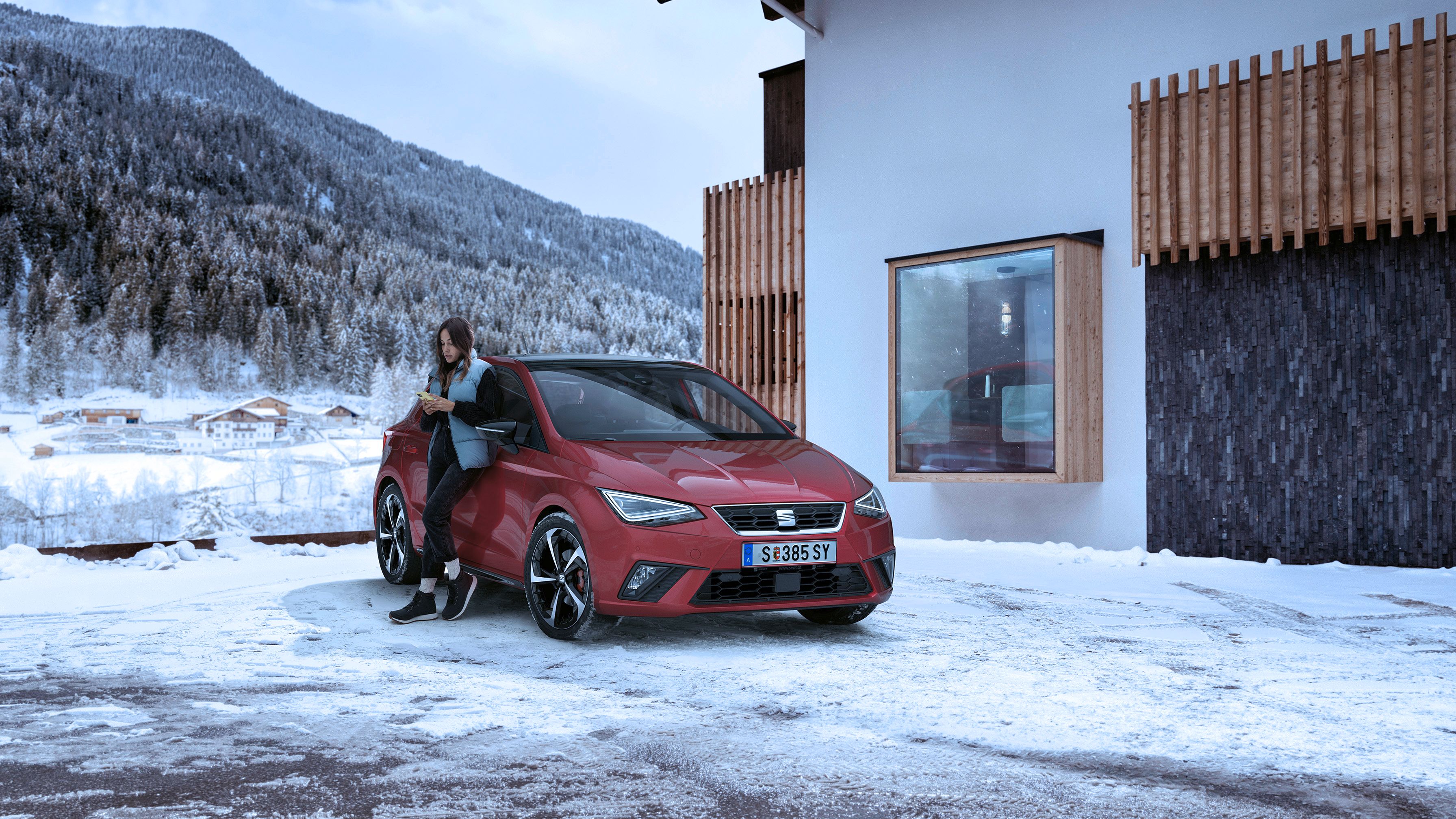 Eine Frau lehnt an einem roten SEAT Ibiza, der in einer verschneiten Landschaft neben einem Haus geparkt ist.