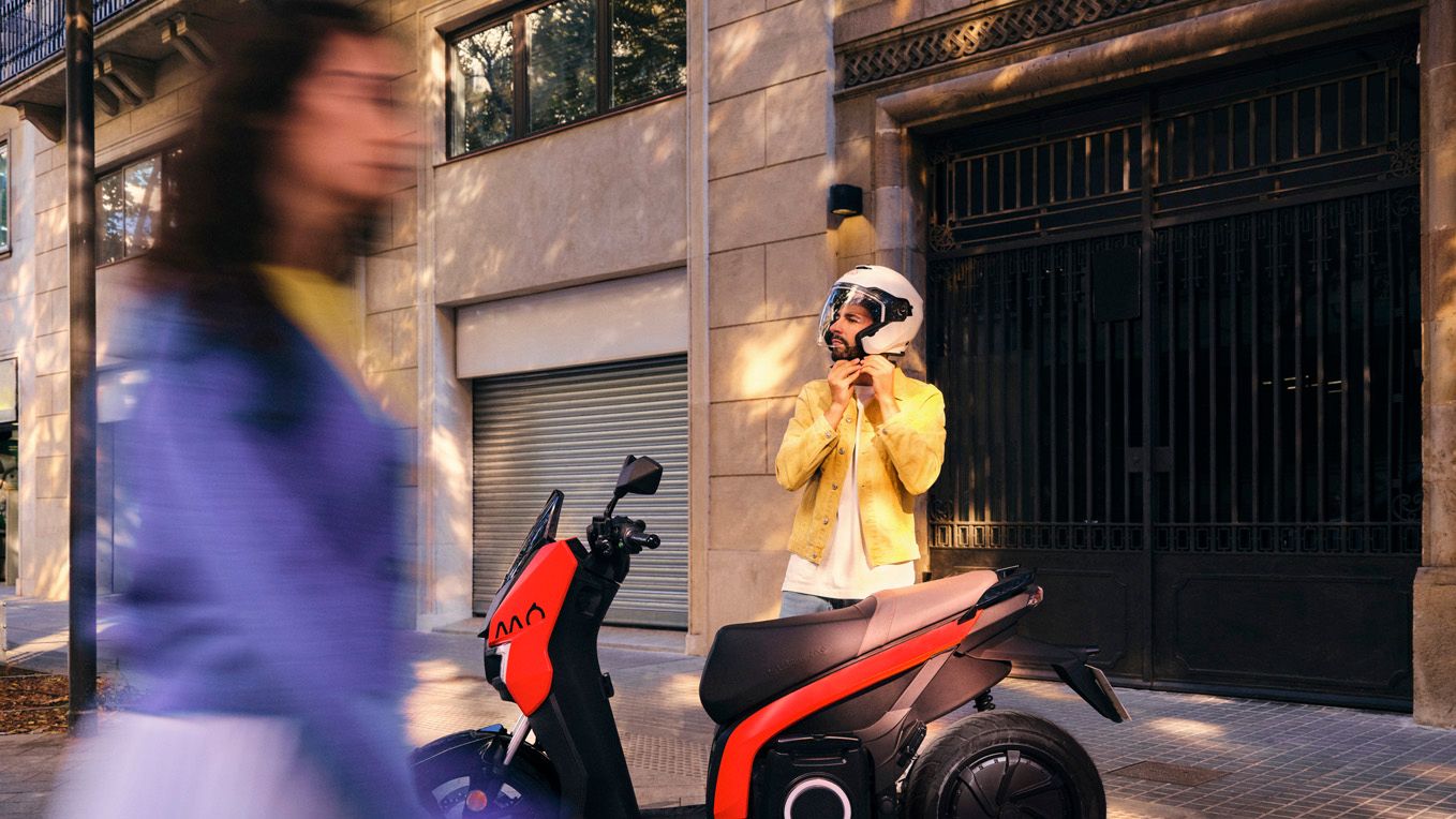 SEAT MO 125 in Emotion Red parkt vor einem Gebäude. Ein Mann steht daneben und setzt sich einen Schutzhelm auf.