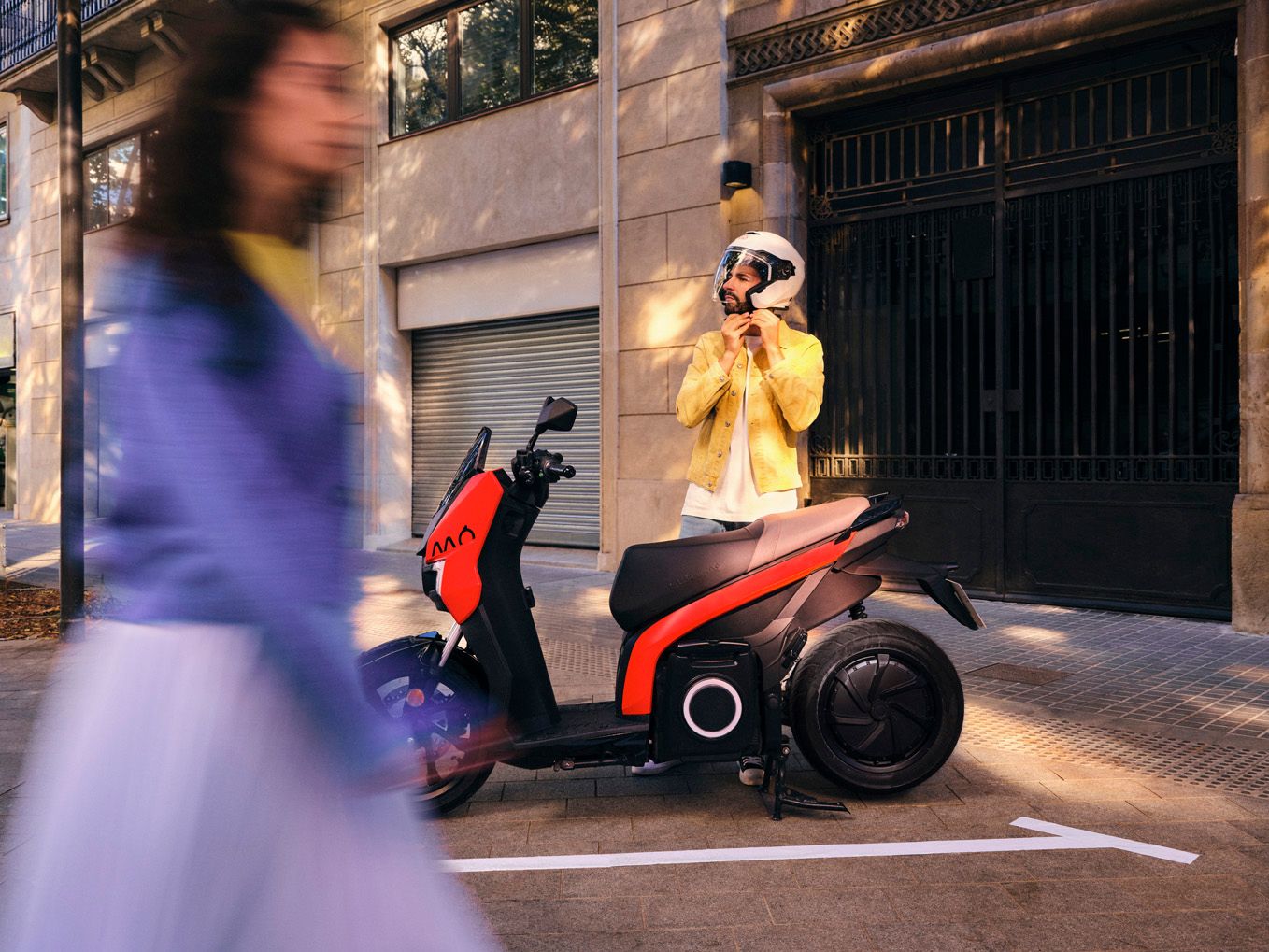 SEAT MO 125 in Emotion Red parkt vor einem Gebäude. Ein Mann steht daneben und setzt sich einen Schutzhelm auf.