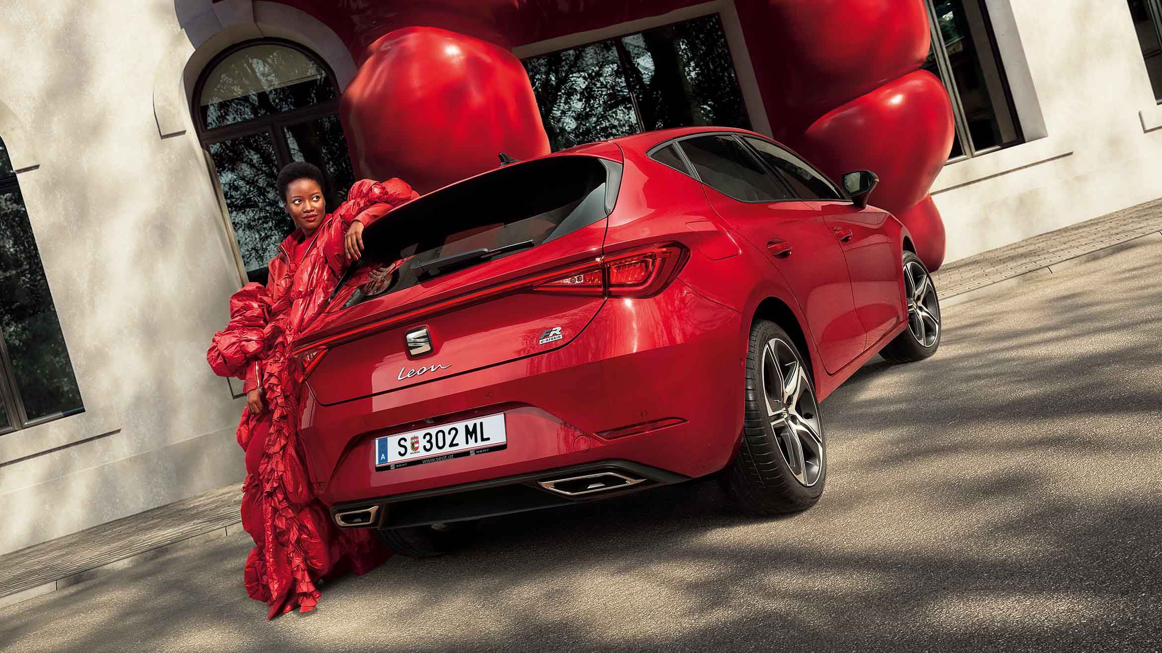 Eine stilvoll in Rot gekleidete Frau lehnt lässig an einem roten SEAT Leon FR e-HYBRID mit modernen LED-Rückleuchten